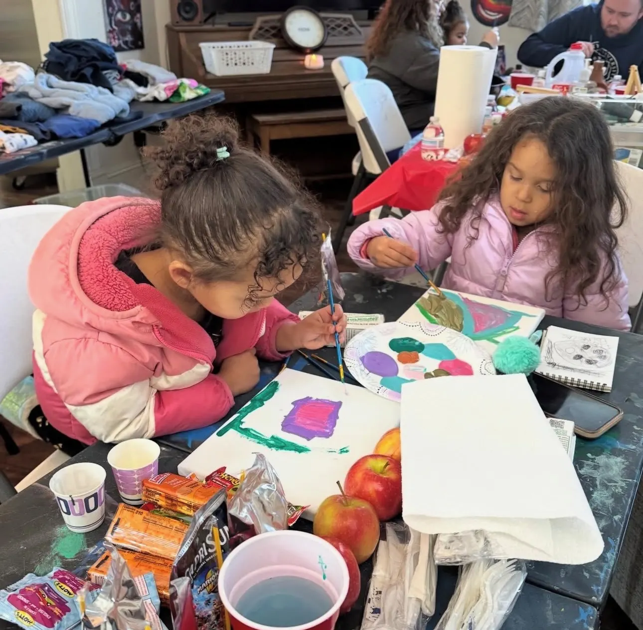 Two little girls in pink jackets paint at a table set with apples, juice and snacks at The Clarity House