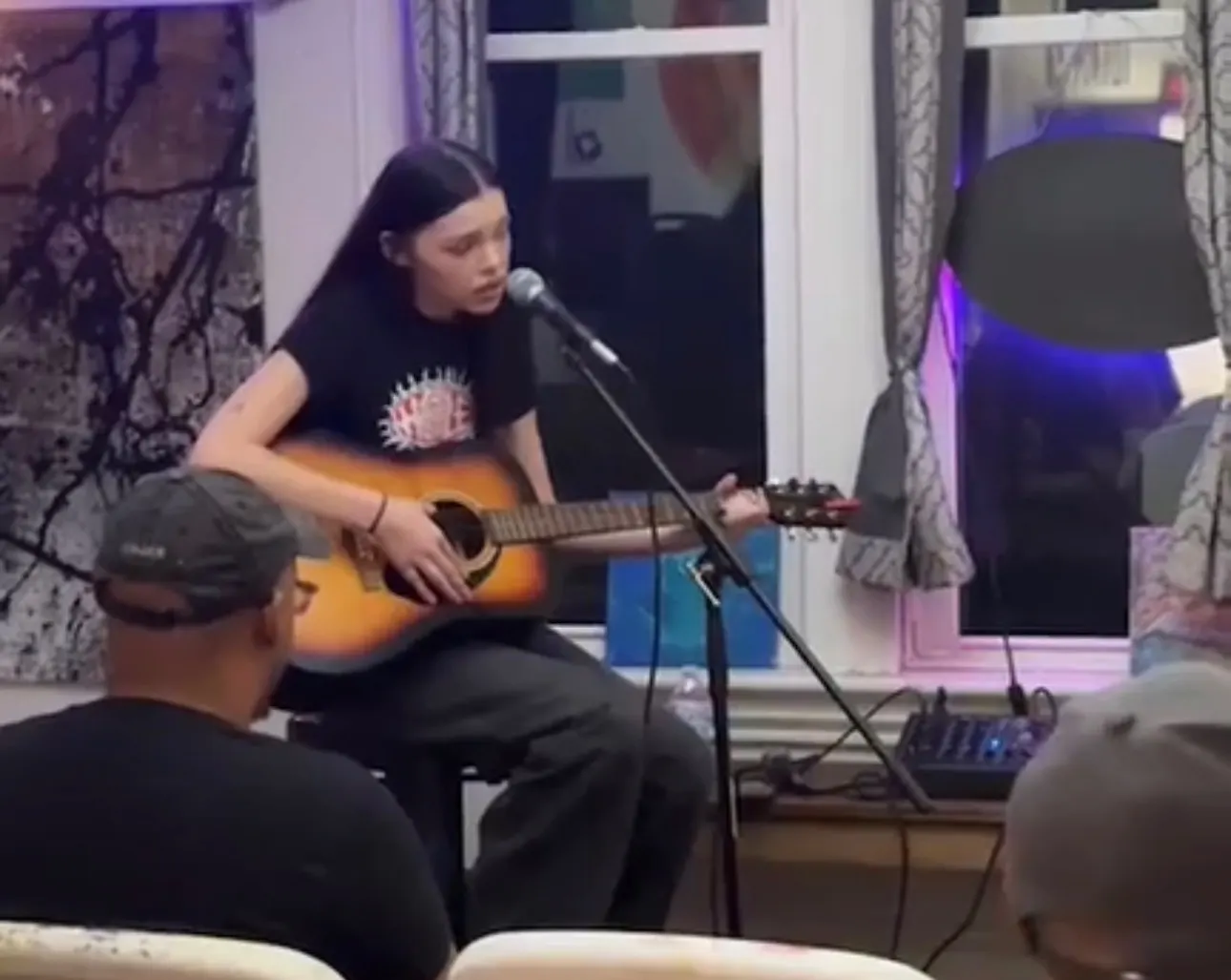A young performer sings and plays acoustic guitar at the mic in the front room