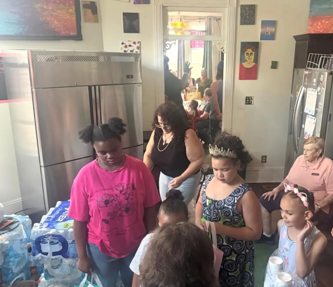 Kids and families in the kitchen at The Clarity House next to stacked cases of drinking water ready to go home with them